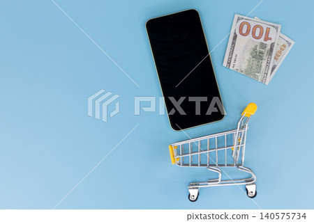 Minimalist scene with smartphone and shopping cart. Flat lay on solid blue background. Modern device beside miniature retail cart. Suggests online shopping or e-commerce concept Minimalist scene with smartphone and shopping cart. Flat lay on solid blue background. Modern device beside miniature retail cart. Suggests online shopping or e-commerce concept 140575734