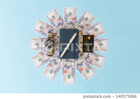 High angle view of a blue notebook and silver pen centered in a sunburst pattern of US dollar bills, accompanied by a luxury wristwatch and credit card on a blue background High angle view of a blue notebook and silver pen centered in a sunburst pattern of US dollar bills, accompanied by a luxury wristwatch and credit card on a blue background 140575806