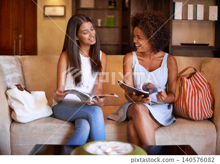 Women, talk and laugh on sofa with magazine for relax, reading or waiting with bags for idea and style. Diverse, friends and smile with article for inspiration, visit or conversation on couch 140576082