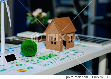 Small model house on energy charts beside a wind turbine and green plant ball on an office desk, showing eco home planning, renewable power, and sustainable real estate. Small model house on energy charts beside a wind turbine and green plant ball on an office desk, showing eco home planning, renewable power, and sustainable real estate. 140576301