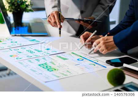 Business team reviewing esg reports and charts on a desk, pointing at data during a meeting in a modern office. Ideal for sustainability, finance and corporate planning themes. Business team reviewing esg reports and charts on a desk, pointing at data during a meeting in a modern office. Ideal for sustainability, finance and corporate planning themes. 140576306