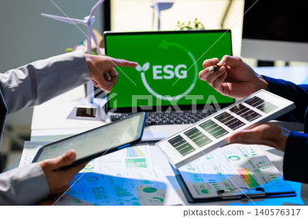 Business team discussing esg strategy at office desk with laptop screen, charts, tablet, solar panel sample and wind turbine models, showing sustainable planning and green investment. 140576317
