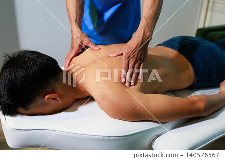 Male patient lying face down on a treatment table while a therapist gives a back massage in a wellness clinic. Close view of hands working on the upper back and shoulders. Male patient lying face down on a treatment table while a therapist gives a back massage in a wellness clinic. Close view of hands working on the upper back and shoulders. 140576367