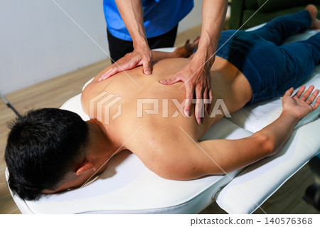 Therapist giving a back massage to a young man lying on a treatment table in a wellness clinic, showing relaxation, body care, pain relief and professional therapy. Therapist giving a back massage to a young man lying on a treatment table in a wellness clinic, showing relaxation, body care, pain relief and professional therapy. 140576368