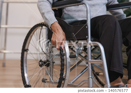 Close view of a senior person sitting in a wheelchair indoors, holding the wheel rim. Image suggests mobility support, recovery, disability care, independence and daily life at home or clinic. Close view of a senior person sitting in a wheelchair indoors, holding the wheel rim. Image suggests mobility support, recovery, disability care, independence and daily life at home or clinic. 140576577