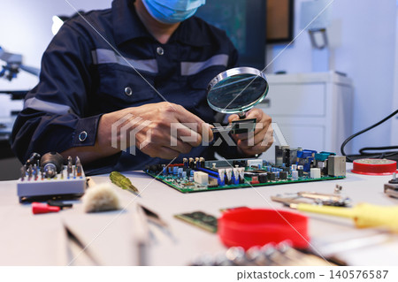 Technician inspecting a circuit board with a magnifying glass at an electronics repair workbench surrounded by tools and components. 140576587