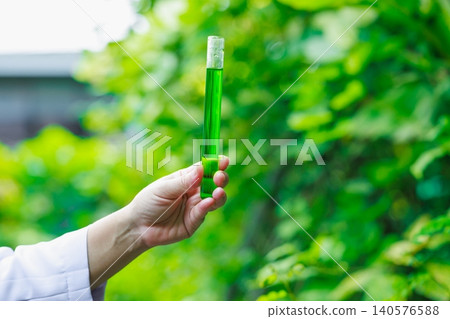 Hand holding a glass test tube filled with green liquid outdoors, with soft blurred leaves in the background, showing science, nature, research, and eco friendly ideas. 140576588