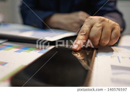 Close-up of a person using a tablet on a desk with printed charts and reports, showing business analysis, data review, planning, and modern office work. Close-up of a person using a tablet on a desk with printed charts and reports, showing business analysis, data review, planning, and modern office work. 140576597