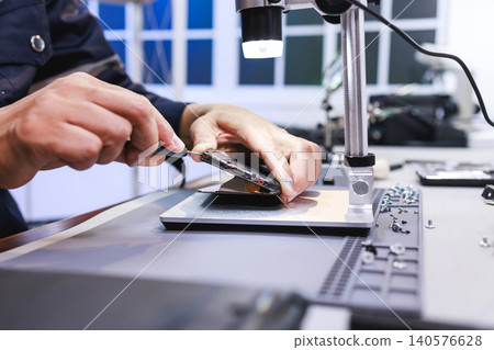 Close-up of a technician repairing a smartphone on a workbench under a small lamp, showing phone maintenance, electronics service, and careful hands-on work in a modern workshop. Close-up of a technician repairing a smartphone on a workbench under a small lamp, showing phone maintenance, electronics service, and careful hands-on work in a modern workshop. 140576628