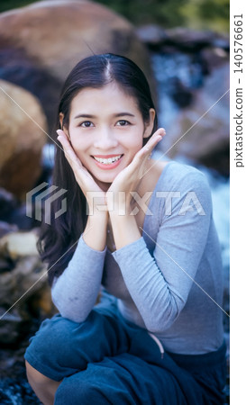 Smiling young woman posing outdoors by rocky stream, hands framing face, natural light, shallow depth of field, travel and lifestyle portrait. 140576661
