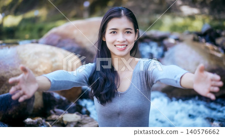 Smiling young woman with arms outstretched inviting a hug outdoors by rocky stream and waterfall, natural light, shallow depth of field, friendly welcoming gesture Smiling young woman with arms outstretched inviting a hug outdoors by rocky stream and waterfall, natural light, shallow depth of field, friendly welcoming gesture 140576662