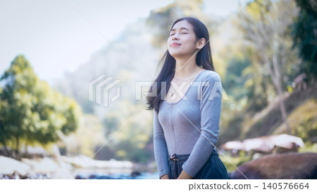 Young woman enjoying fresh air outdoors by a riverside, eyes closed, calm smile, soft morning light with blurred forest mountains in background Young woman enjoying fresh air outdoors by a riverside, eyes closed, calm smile, soft morning light with blurred forest mountains in background 140576664