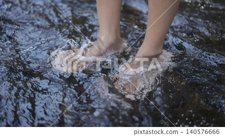 Young woman with arms outstretched enjoying fresh air by a river in a green forest, serene outdoor wellness moment in soft daylight. 140576666