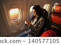 Happy young adult Asian woman using smartphone inside airplane cabin next to window at sunset 140577105
