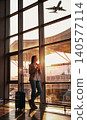 Smiling young adult Caucasian woman holding smartphone next to suitcase at airport terminal 140577114