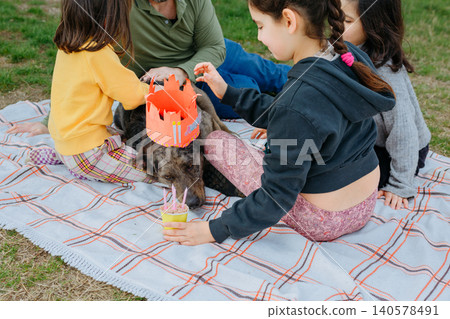 Children Celebrating Dog Birthday Picnic Outdoors with Homemade Crown and Cake Children Celebrating Dog Birthday Picnic Outdoors with Homemade Crown and Cake 140578491
