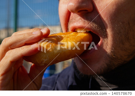 Man eating a fried pirozhok with potato filling. Traditional food snack outdoors. Close-up view of hungry guy enjoying a rustic deep-fried dough pastry. Russian cuisine street food. Man eating a fried pirozhok with potato filling. Traditional food snack outdoors. Close-up view of hungry guy enjoying a rustic deep-fried dough pastry. Russian cuisine street food. 140578670