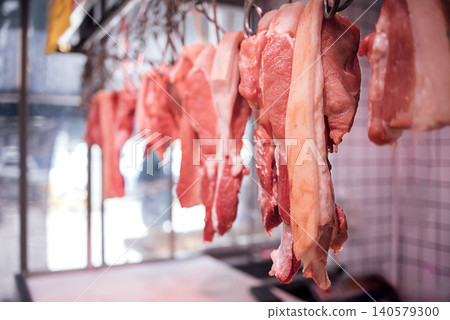 Fresh cuts of pork hanging from hooks in a butcher shop, showcasing various meat textures and colors against a blurred background of the market environment Fresh cuts of pork hanging from hooks in a butcher shop, showcasing various meat textures and colors against a blurred background of the market environment 140579300