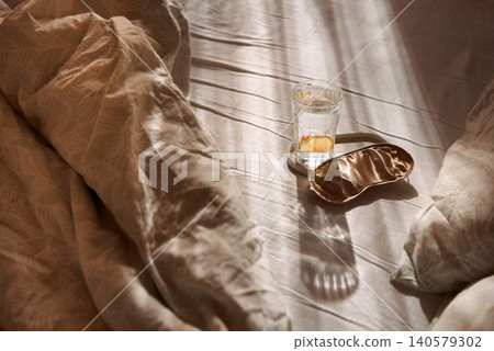 Cozy bedroom scene featuring a glass of water, a silk sleep mask, and crumpled bedding illuminated by soft sunlight streaming through a window Cozy bedroom scene featuring a glass of water, a silk sleep mask, and crumpled bedding illuminated by soft sunlight streaming through a window 140579302