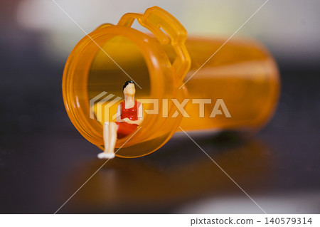 Miniature figure of a woman in a red dress sitting inside an orange prescription pill bottle on a blurred dark background Miniature figure of a woman in a red dress sitting inside an orange prescription pill bottle on a blurred dark background 140579314