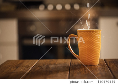 Orange ceramic mug with textured surface filled with hot water, steam rising, placed on wooden table in a kitchen setting with blurred background elements Orange ceramic mug with textured surface filled with hot water, steam rising, placed on wooden table in a kitchen setting with blurred background elements 140579315