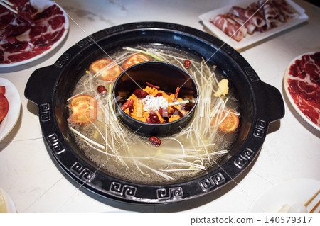 Hot pot dish with broth, vegetables, and spices served in a black pot, surrounded by plates of sliced meat and fresh ingredients on a dining table 140579317