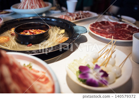Hot pot dining scene featuring a black pot filled with broth, surrounded by various raw ingredients including sliced meat, vegetables, and skewered items on a table Hot pot dining scene featuring a black pot filled with broth, surrounded by various raw ingredients including sliced meat, vegetables, and skewered items on a table 140579372