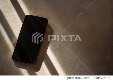 Smartphone lying on a textured surface with soft shadows and light patterns creating a visually appealing composition in a minimalist setting Smartphone lying on a textured surface with soft shadows and light patterns creating a visually appealing composition in a minimalist setting 140579406
