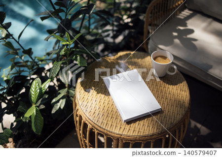 White notebook placed on a round wicker table beside a cup of coffee and green plants near a swimming pool in a serene outdoor setting White notebook placed on a round wicker table beside a cup of coffee and green plants near a swimming pool in a serene outdoor setting 140579407