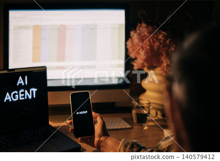 Person holding smartphone displaying "AI AGENT" text while seated at desk with laptop and computer monitor showing spreadsheet and decorative flowers in the background Person holding smartphone displaying "AI AGENT" text while seated at desk with laptop and computer monitor showing spreadsheet and decorative flowers in the background 140579412