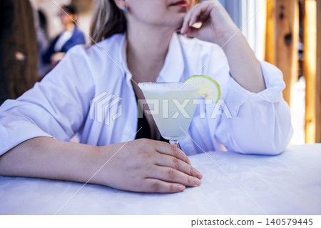Woman with long hair in a white shirt is resting her chin on her hand while holding a cocktail glass with lime slice on a table, enjoying a refreshing drink in a relaxed setting Woman with long hair in a white shirt is resting her chin on her hand while holding a cocktail glass with lime slice on a table, enjoying a refreshing drink in a relaxed setting 140579445