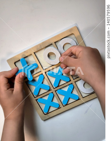 A child is arranging wooden tic-tac-toe pieces on a white table. A child is arranging wooden tic-tac-toe pieces on a white table. 140579596