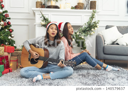 Happy Asian Woman sitting Christmas gift box under xmas tree playing acoustic guitar together. Two Women smile talk sitting decor christmas ball tree xmas holiday winter festival in living room Happy Asian Woman sitting Christmas gift box under xmas tree playing acoustic guitar together. Two Women smile talk sitting decor christmas ball tree xmas holiday winter festival in living room 140579637