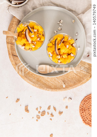 A top view of two glass parfaits filled with diced mango and coconut flakes on a round wooden board and grey plate surrounded by rustic linens and crumbs for a healthy breakfast concept A top view of two glass parfaits filled with diced mango and coconut flakes on a round wooden board and grey plate surrounded by rustic linens and crumbs for a healthy breakfast concept 140579749