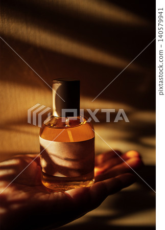 Glass perfume bottle with a golden liquid held in a hand, soft shadows and warm light creating an elegant atmosphere against a textured background Glass perfume bottle with a golden liquid held in a hand, soft shadows and warm light creating an elegant atmosphere against a textured background 140579941