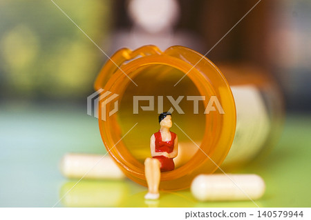 Miniature figure of a woman in a red dress sitting inside an overturned prescription pill bottle with capsules scattered around on a colorful surface Miniature figure of a woman in a red dress sitting inside an overturned prescription pill bottle with capsules scattered around on a colorful surface 140579944
