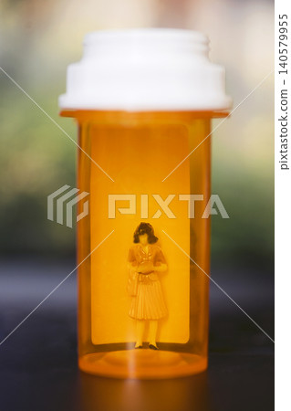 Orange prescription bottle containing a small figurine of a woman dressed in a yellow dress, with a blurred background creating a soft focus effect Orange prescription bottle containing a small figurine of a woman dressed in a yellow dress, with a blurred background creating a soft focus effect 140579955