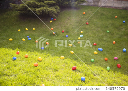 Colorful plastic Easter eggs scattered across a green lawn in a backyard setting, surrounded by shrubs and trees under bright sunlight Colorful plastic Easter eggs scattered across a green lawn in a backyard setting, surrounded by shrubs and trees under bright sunlight 140579970