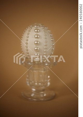 Decorative white egg adorned with pearls displayed on a clear glass pedestal against a warm brown background, suitable for festive or elegant occasions Decorative white egg adorned with pearls displayed on a clear glass pedestal against a warm brown background, suitable for festive or elegant occasions 140580002