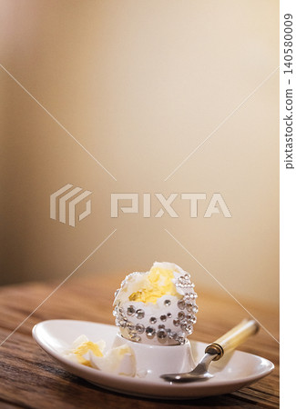 Decorative dessert with whipped cream and silver embellishments served on a white plate, accompanied by a small spoon on a wooden table Decorative dessert with whipped cream and silver embellishments served on a white plate, accompanied by a small spoon on a wooden table 140580009
