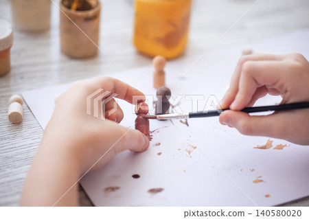 Child's hand painting wooden figurines with a brush on a white sheet of paper, jars of paint in the background, creative art project in a bright indoor space Child's hand painting wooden figurines with a brush on a white sheet of paper, jars of paint in the background, creative art project in a bright indoor space 140580020