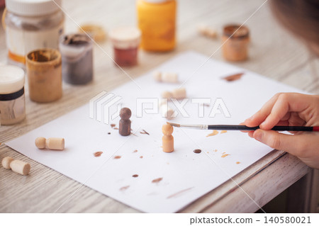 Hand of a person painting wooden figurines with a brush on a white sheet, surrounded by jars of paint in various colors on a wooden table Hand of a person painting wooden figurines with a brush on a white sheet, surrounded by jars of paint in various colors on a wooden table 140580021