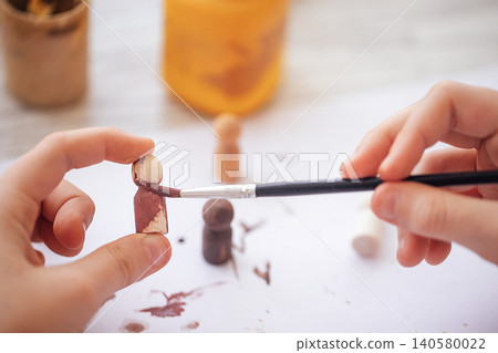 Child's hands painting wooden figurines with a brush, jars of paint visible in the background on a light-colored table surface Child's hands painting wooden figurines with a brush, jars of paint visible in the background on a light-colored table surface 140580022