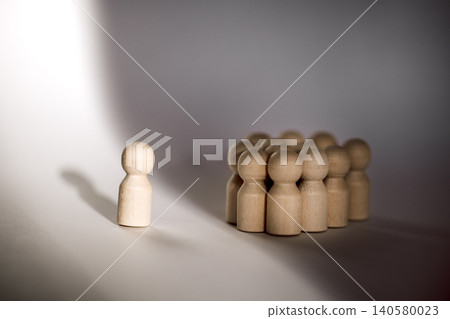 Wooden figurine standing alone in front of a group of ten wooden figures, casting shadows on a light background, representing individuality and social dynamics Wooden figurine standing alone in front of a group of ten wooden figures, casting shadows on a light background, representing individuality and social dynamics 140580023