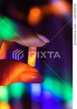 Close-up of a hand holding a translucent capsule against a colorful blurred background with vibrant lighting effects and reflections Close-up of a hand holding a translucent capsule against a colorful blurred background with vibrant lighting effects and reflections 140580030
