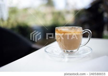 Glass cup of latte coffee with creamy foam on top placed on a clear saucer, sitting on a white table with blurred greenery in the background Glass cup of latte coffee with creamy foam on top placed on a clear saucer, sitting on a white table with blurred greenery in the background 140580033