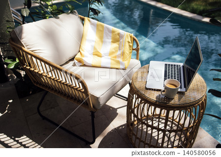 Cozy outdoor seating area with a rattan chair, striped blanket, laptop, and coffee cup beside a swimming pool surrounded by lush greenery Cozy outdoor seating area with a rattan chair, striped blanket, laptop, and coffee cup beside a swimming pool surrounded by lush greenery 140580056