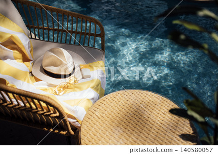 Woven rattan chair with yellow striped towel, sun hat, and sunglasses beside a sparkling blue swimming pool in a tropical outdoor setting 140580057