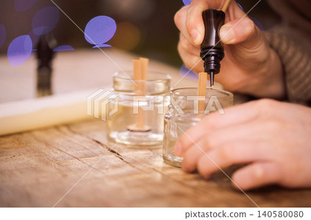 Hands of a person carefully pouring wax into small glass jars with wooden wicks, creating homemade candles, showcasing the art of candle making and craftsmanship in a cozy environment 140580080