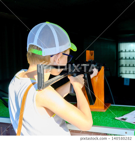 rear view of a boy in a baseball cap and glasses shooting from an automatic rifle on a wooden stand in a range rear view of a boy in a baseball cap and glasses shooting from an automatic rifle on a wooden stand in a range 140580282